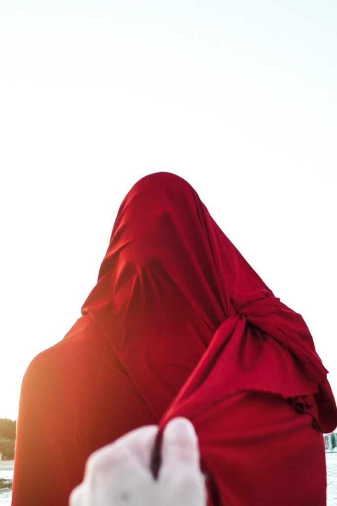 Faceless person pulling edge of red cloth wrapping faceless person standing against cloudless blue sky